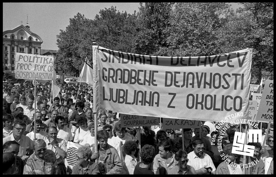 Množica ljudi, v rokah držijo napise. Na enem piše: Sindikat delavcev gradbene dejavnosti Ljubljana z okolico.