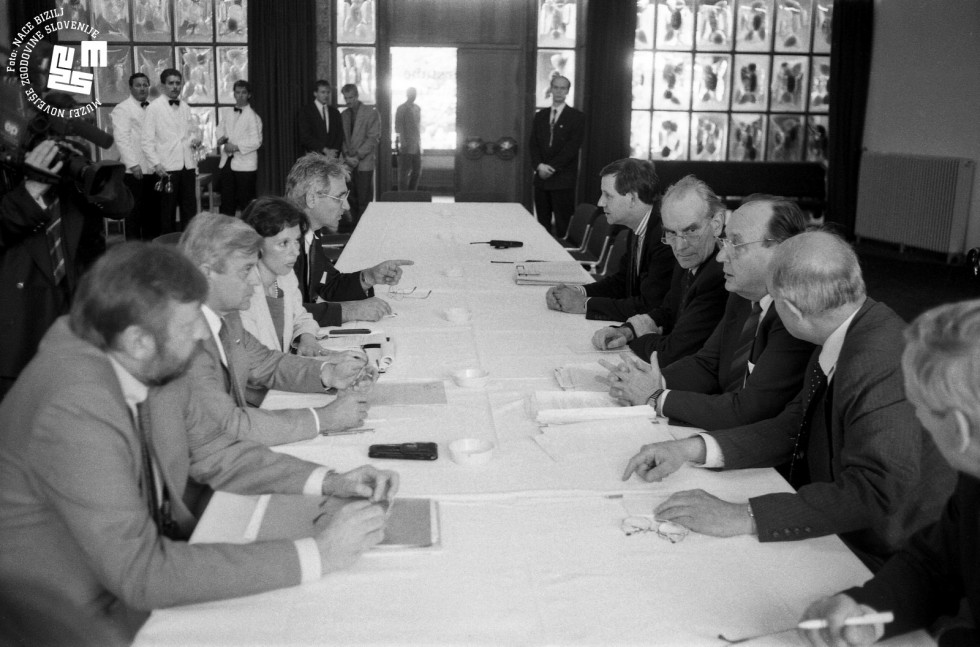 Hans Dietrich Genscher sedi za mizo in govori. Poleg njega sedijo še štirje moški. Nasproti sedijo Dimitrij Rupe, Milan Kučan, prevajalka in moški.