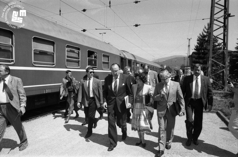 Vlak na levi strani, po peronu hodi nemška in slovenska delegacija., med njimi Hans Dietrich Genscher, poleg prevajalka, zraven Milan Kučan, zadaj Dimitrij Rupel.