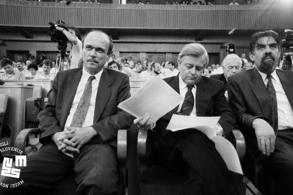 Janez Drnovšek, Milan Kučan, ki gleda papirje, Ciril Zlobec sedijo v skupščini. V ozadju ostali člani skupščine.