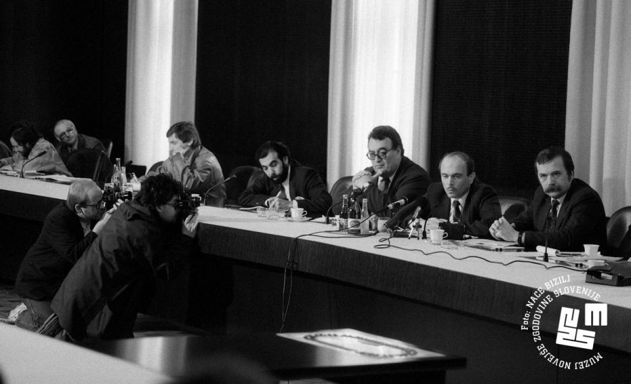 Janez Drnovšek sedi za mizo in govori na tiskovni konferenci. Poleg sedijo ostali govorci.