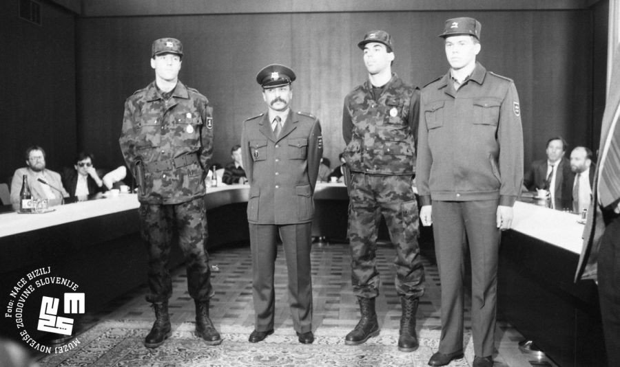 V sejni sobi sedijo ljudje, štiri osebe v uniformah stojijo v polkrogu.