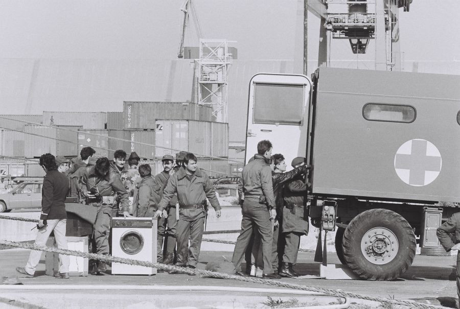 Vojaki v pristanišču stojijo pri tovornjaku, ki ima odprta zadnja vrata. Nekateri vojaki gledajo v njega. Na tleh je pralni stroj in dva večja paketa.
