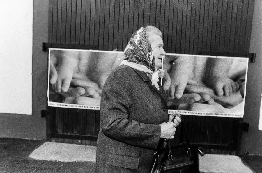 Stara gospa z ruto na glavi gre mimo plakatov, ki vabijo na plebiscit.