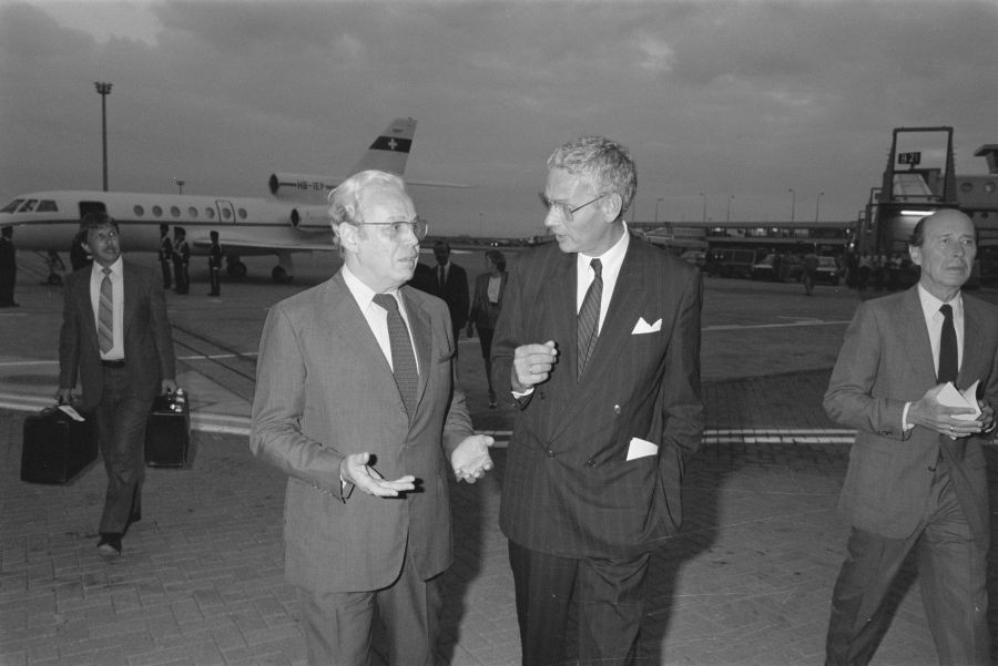 Perez De Cuellar in Hans van den Broek se pogovarjata na letališču. V ozadju letalo, levo hodi moški s kovčki, desno gre mimo moški.