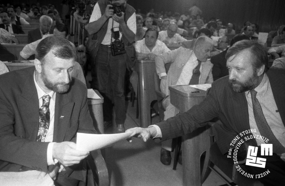 Dušan Plut in Dimitrij Rupel sedita v skupščini. Plut drži v rokah dokument, Rupel mu nekaj kaže na njem. V ozadju kamerman, ostali člani skupščine sedijo za njima.
