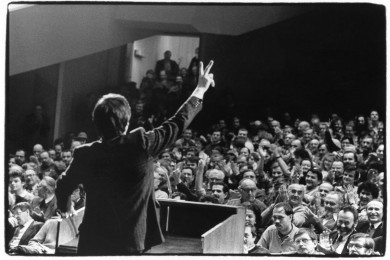 Moški na govorniškem odru drži roko navzgor s prsti v obliki črke V. Pogled na polno dvorano ljudi, ki ploskajo.