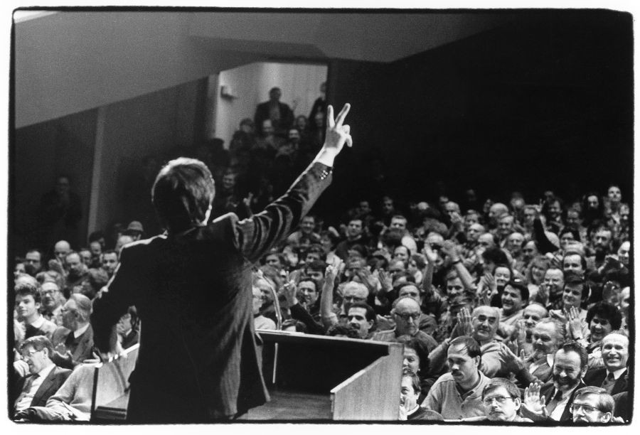 Moški na govorniškem odru drži roko navzgor s prsti v obliki črke V. Pogled na polno dvorano ljudi, ki ploskajo.