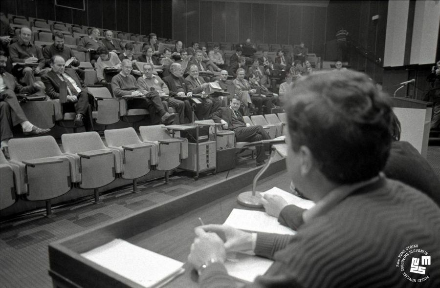 Člani sedijo na tribuni, oseba sedi na govorniškem odru.