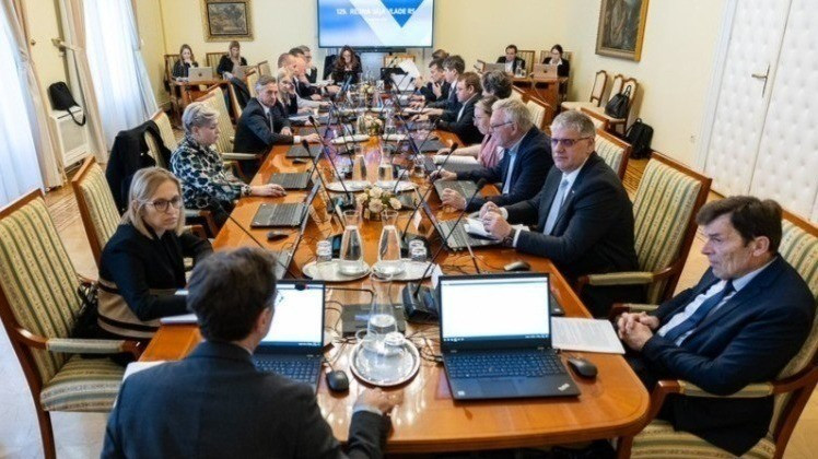 Government officials seated around a long wooden conference table during a formal cabinet meeting, each working on laptops, with documents and water pitchers on the table.