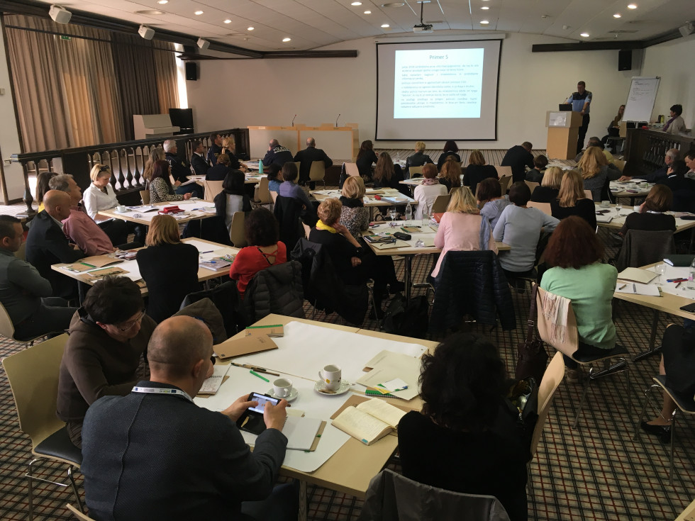 Udeleženci sedijo za mizami v skupinah, govornik na odru