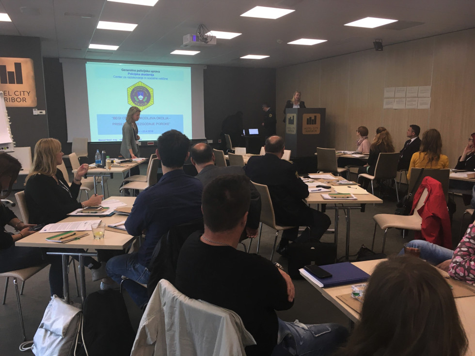 Udeleženci sedijo za mizami po skupinah, govorka stoji na odru