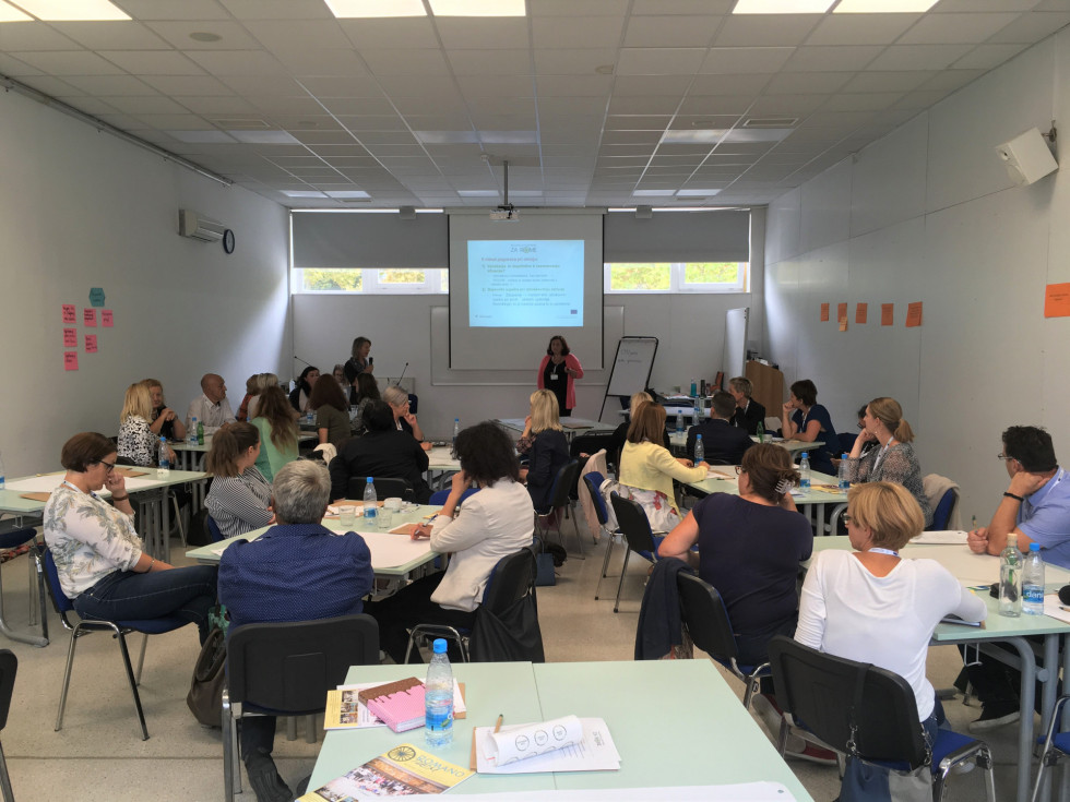 Udeleženci sedijo za mizami v skupinah, predavateljica stoji pred tablo