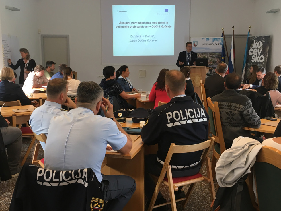 Udeleženci sedijo za mizami, govorec za govorniškim pultom