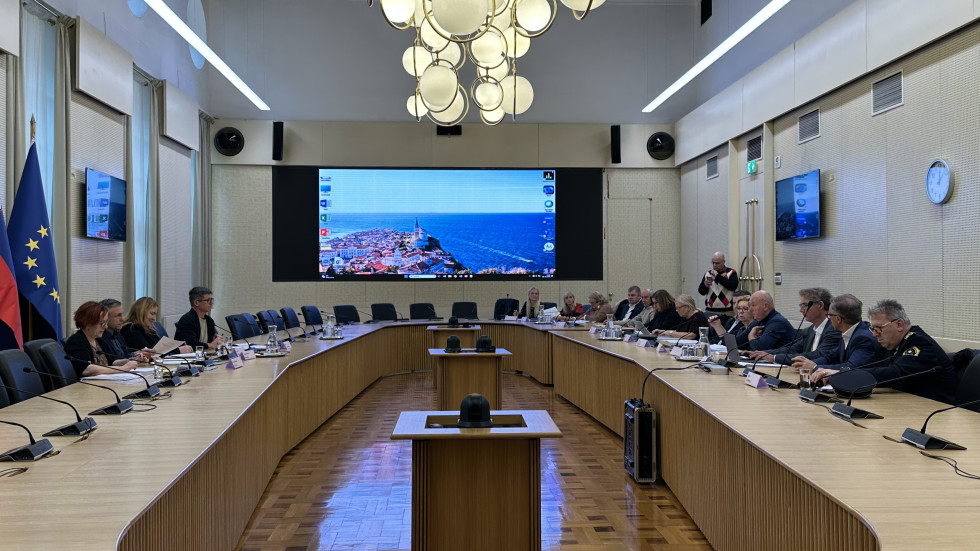 Udeleženci seje sedijo v sejni dvorani, v ozadju projekcija