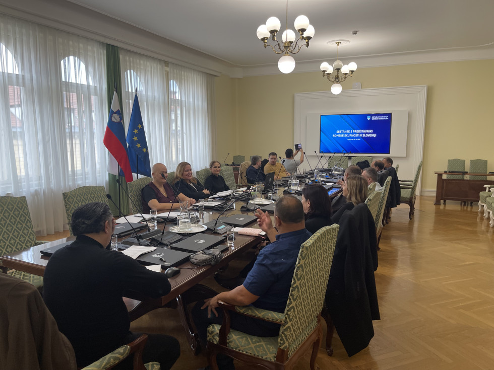 Predstavniki urada in predstavniki romske skupnosti sedijo za mizo. Na steni velik ekran s predstavitvijo.