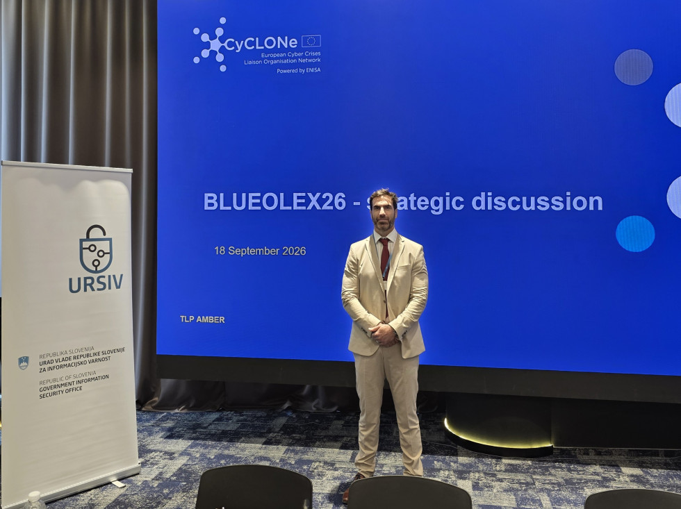 Moški stoji pred velikim modrim ekranom z napisom: BlueOLEx26 - strategic discussion.