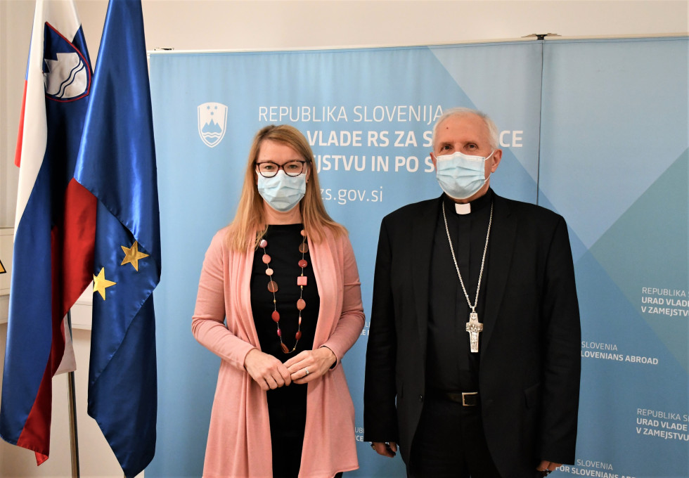 Ministrica in nadškof pred plakatom urada, na desni slovenska in evropska zastava.