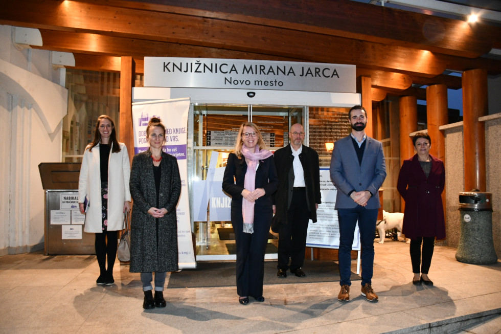 Ministrica in direktor knjižnjice in ostali stojijo pred vrati knjižnjice, zgoraj napis Knjižnica Mirana Jarca Novo Mesto.