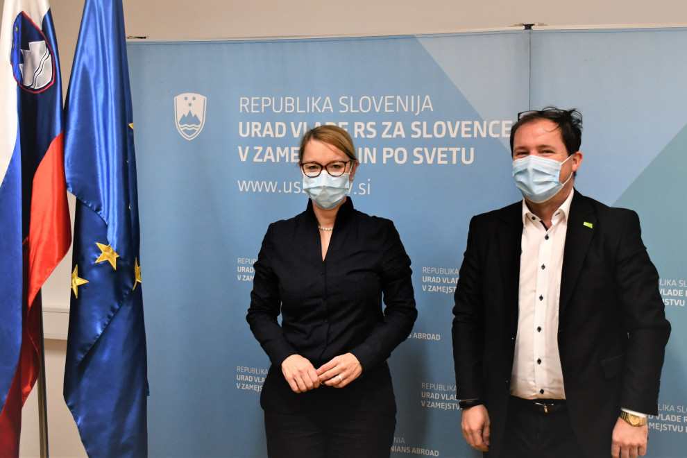 Ministrica in diretor pred plakatom urada, ob strani zastavi.