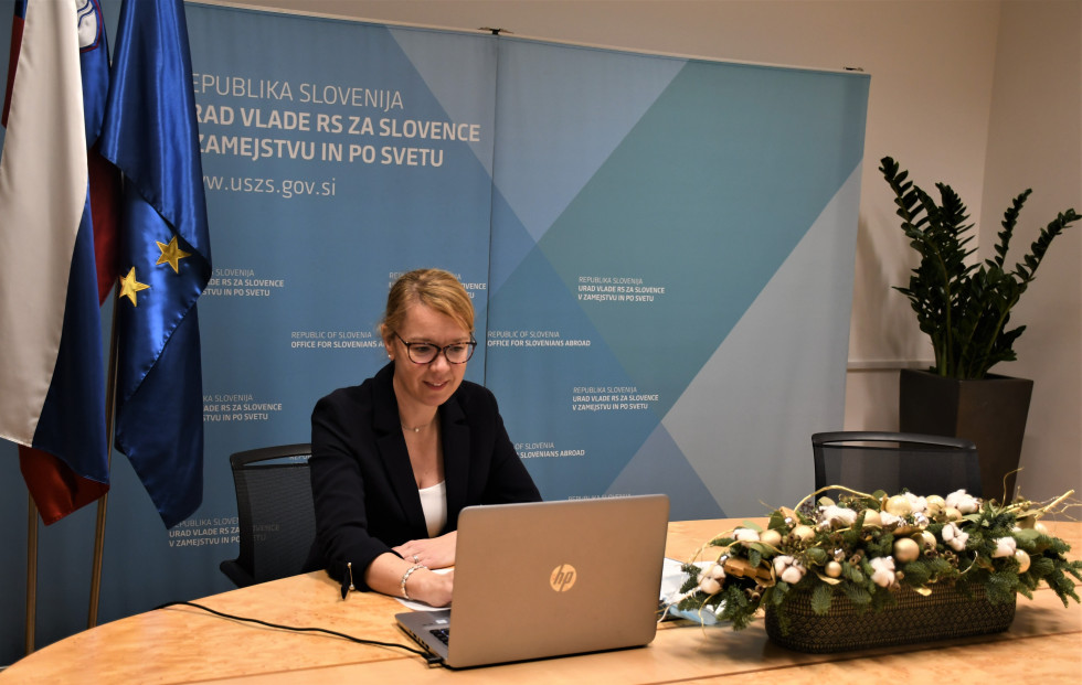 Ministrica sedi za mizo, na desni strani slovenska in evropska zastva. Pred njo računalnik, v ozadju napis Urad,