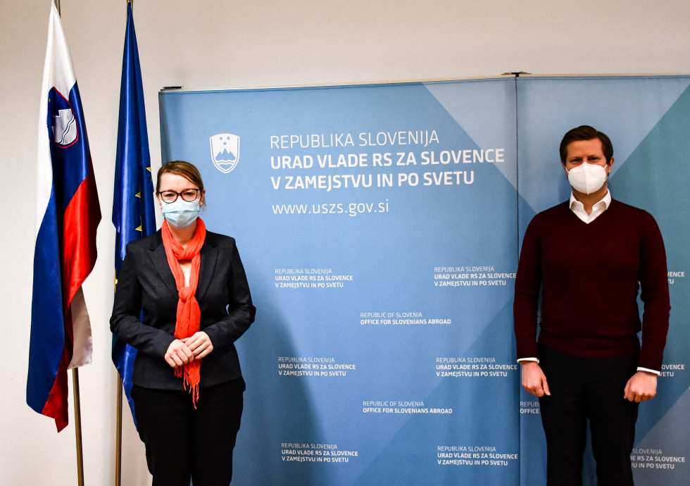 Stojita pred panojem z napisom Urad Vlade RS za Slovence v zamejstvu in po svetu, na desni strani slovenska in evropska zastava.