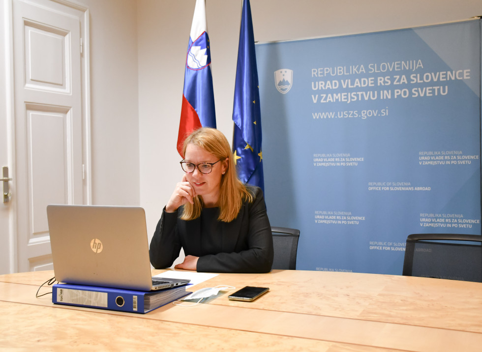Za računalnikom, sedi, za njo slovenska zastava in zastava EU in stena z napisi Urad za Slovence v zamejstvu in po svetu.