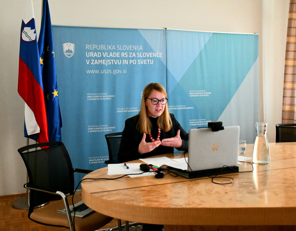 Med pogovorom, ministrica sedi za mizo, pred njo računalnik, za njo slovensk zastava in zastava EU.
