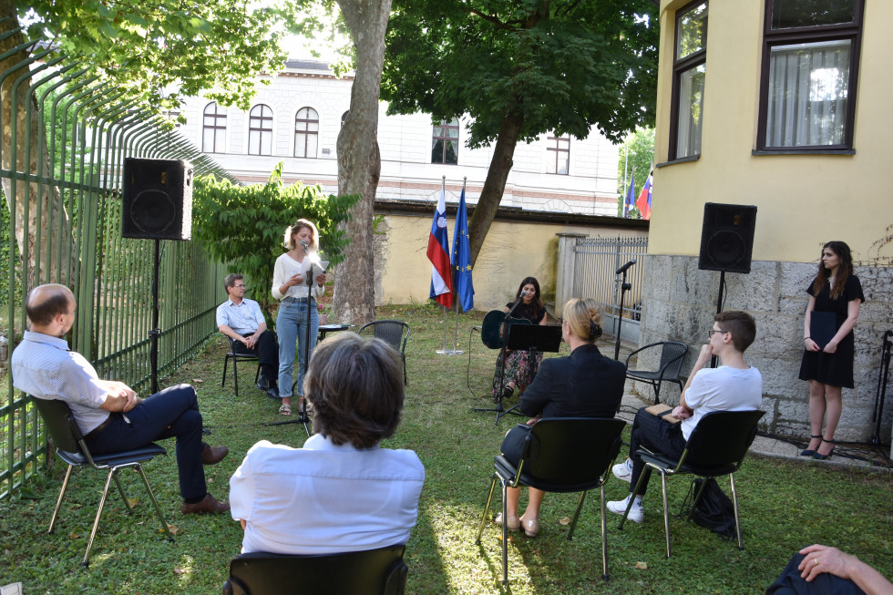 Na vrtu urada, Verena pri mikrofunu, v hrbet obiskovalci, Vincenc sedi, Nadia sedi. 