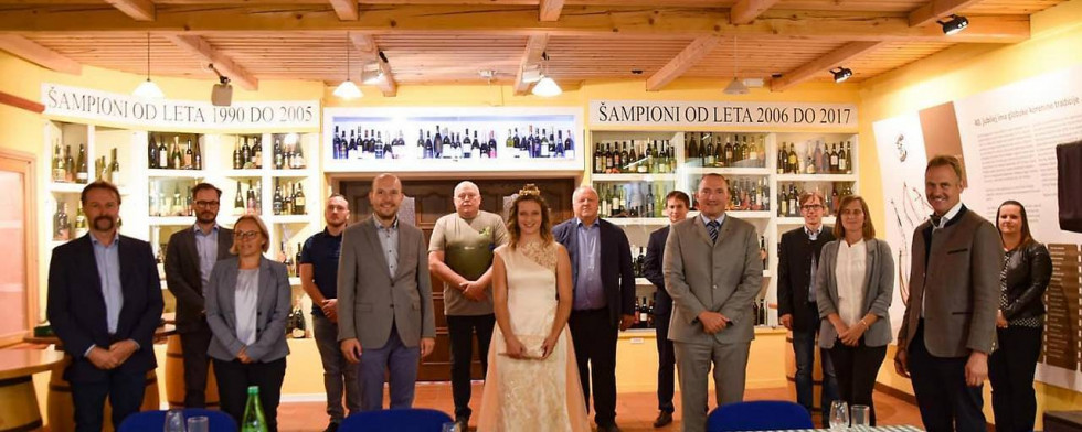 Udeleženci srečanja stojijo, na sredini vinska kraljica, v ozadju na policah steklenice vina. 
