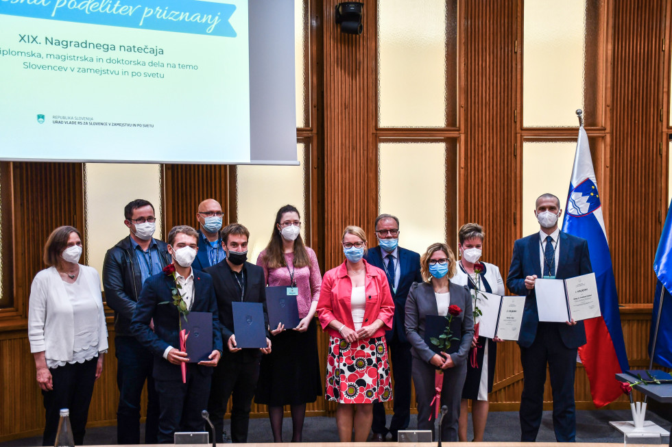 Ministrica, nagrajenci in člani komisije stojijo, na desni strani slovenska in evropska zastava. Nagrajenci imajo v rokah prizananja in vrtnice.