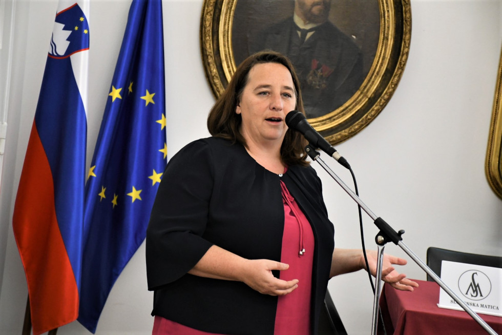 Vesna Humar, na njeni desni strani v ozadju slovenska in evropska zastava. Državna sekretarka govori na mikrofon.
