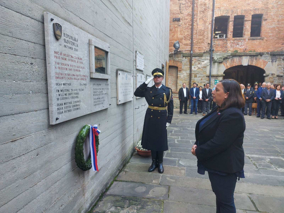 Džravna sekretarka položila venec v Rižani. Stoji pred spominsko ploščo, pod njo lovorjev venec.