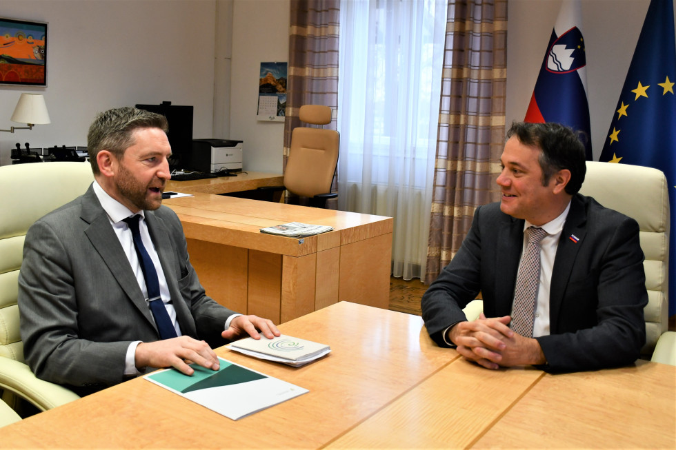 Adrian Farrell in Matej Arčon sedita za mizo, v ozadju slovenska in evropska zastava.