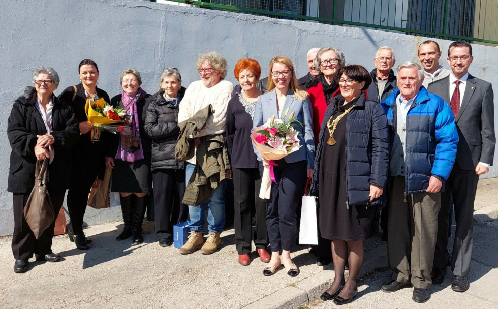 Ministrica z rojaki zunaj na soncu, v rokah drži šopek rož.