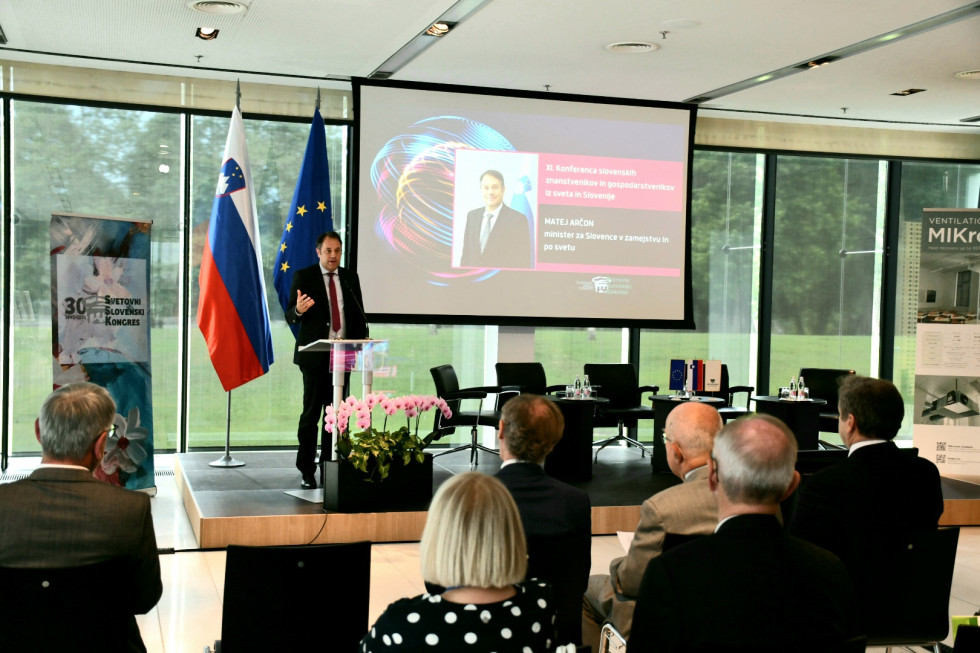 Minister za govornico, na strani slovenska in evropska zastava, na drugi strani ekran, v dvorani obiskovalci.