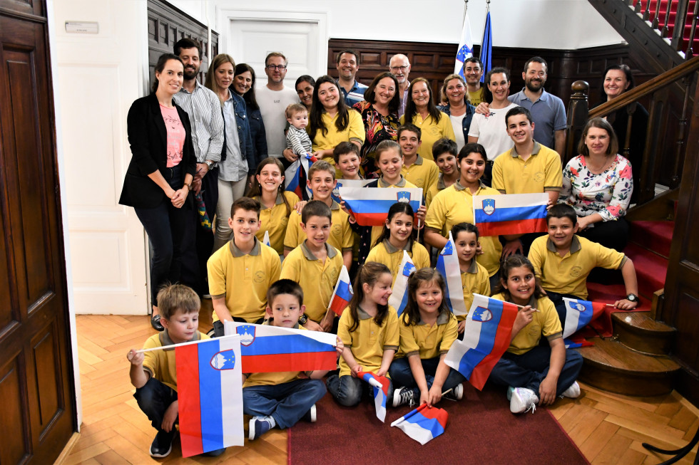 Državna sekretarka s člani otroškega zbora Škrjančki, njihovo babico, starši in sodelavci urada. Škrjančki imajo v rokah slovenske zatave in oblečene rumene majice.