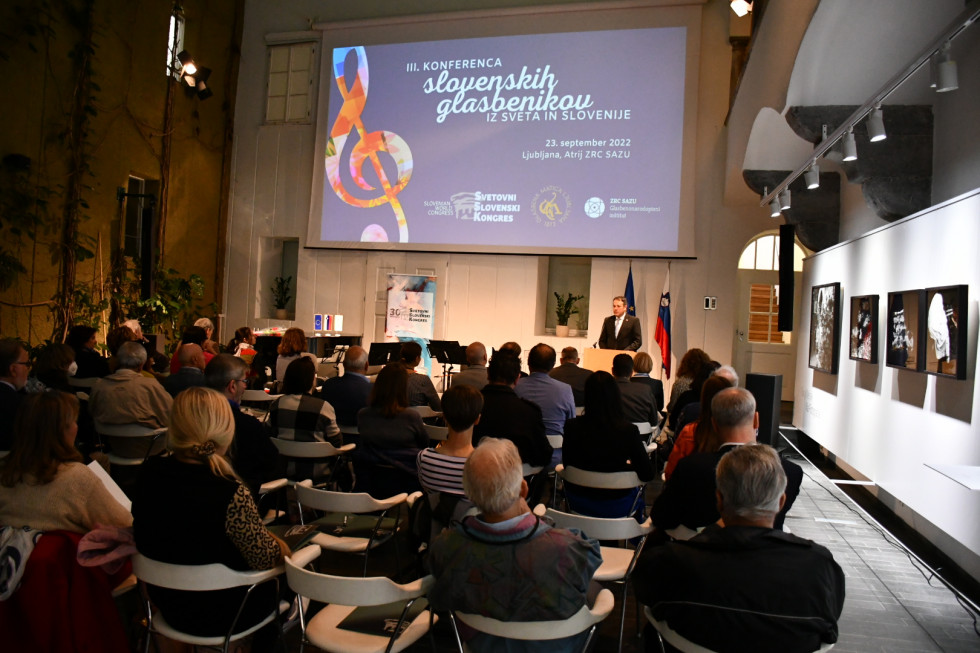 Minister Matej Arčon med govorom. Posnetek v hrbet udeležencem, nad ministrom zaslon z napisom III. konferenca slovenskih glasbenikov iz sveta in Slovenije