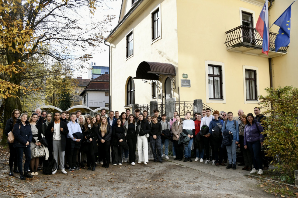 Mladi z ministrom pred stavbo urada, na porčelju visita zastavi.