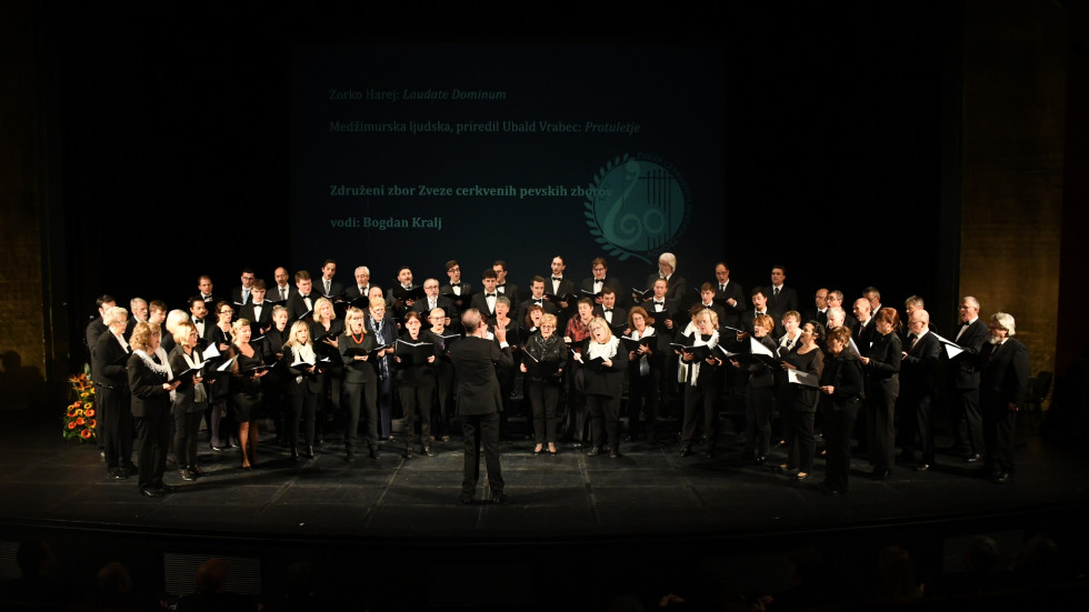 rkvenih pevskih zborov med nastopom na odru, v ozadju predstavitev z napisanim imenom in repertuarjem. 