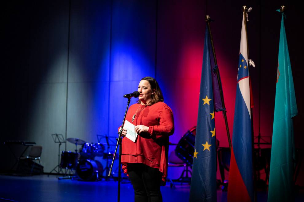 Državna sekretarka na odru za mikrofonom, za njo slovenska, evropska in novogoriška zastava.