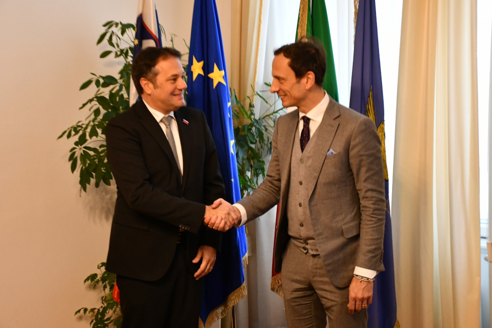 Matej Arčon in Massimiliano Fedriga se rokujeta. Za njima zastave (slovenska, evropska, italijanska, zastava Furlanije-Julijske krajine).
