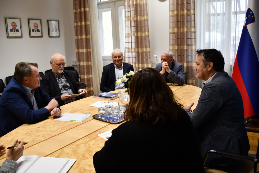 Udeleženci sestanka. Sedijo za mizo, minister in državna sekretarka fotografirana v hrbet, na desni zastave.