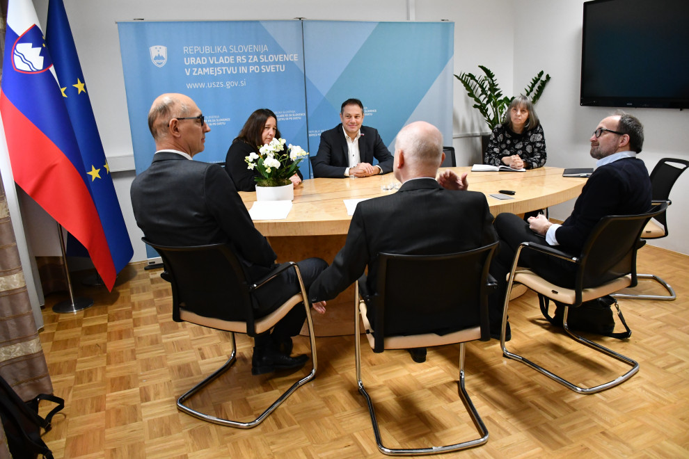 Udeleženci sestanka sedijo za mizo, v ozadju pano urada, na desni slovenska in evropska zastava.