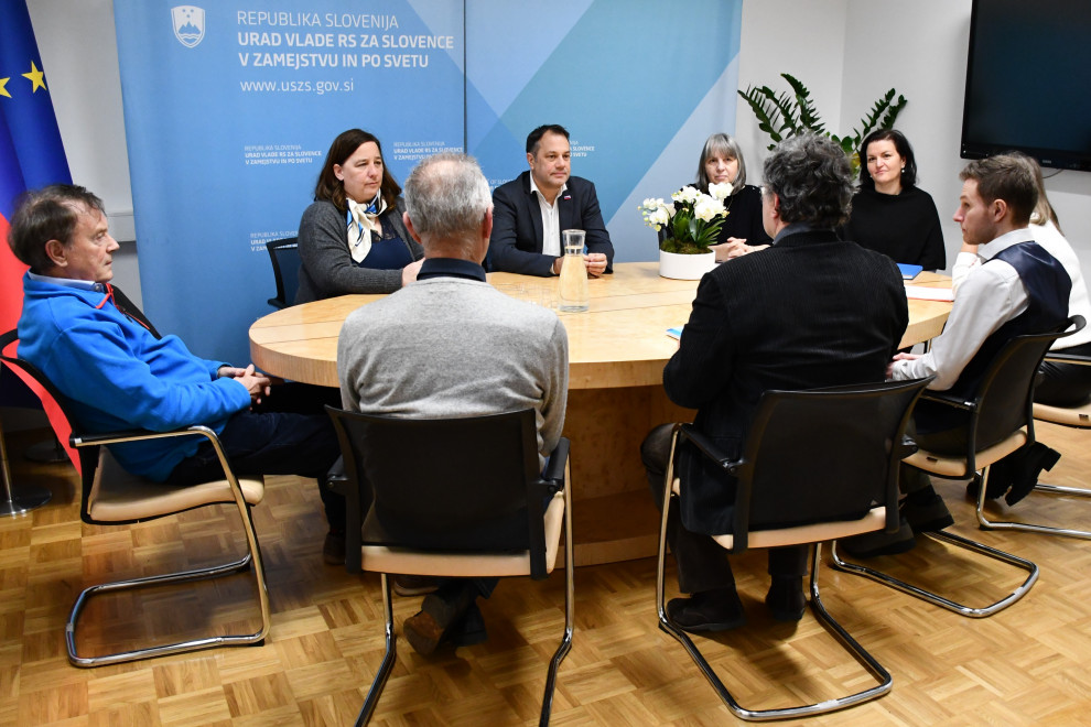 Udeleženci sestanka za mizo, v ozadju plakat z logotipi urada, na desni slovensa in evropska zastava.