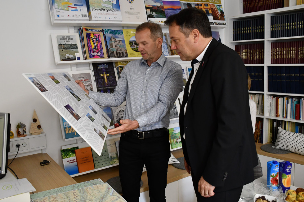 Lenart Rihar in Matej Arčon gledata v plakat, na katerem je predstavljen zemljevid s slovenskimi imeni zamejskih krajev v vseh štirih sosednjih državah. Zemljevid v rokah drži Leart Rihar, minister pa gleda.