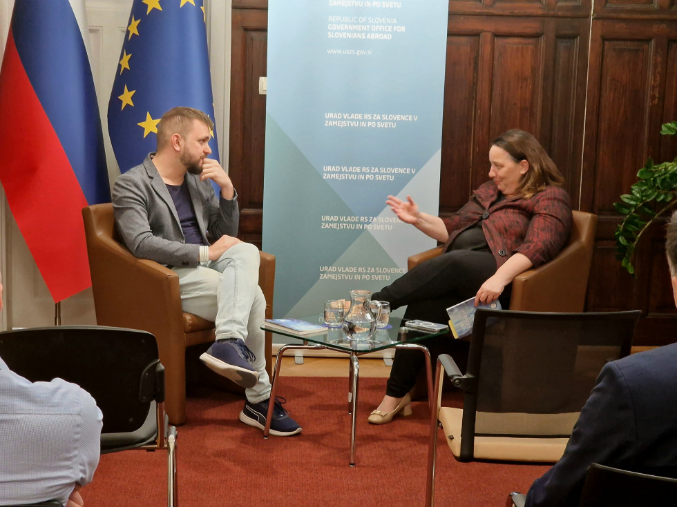 Alex Kama Devetak in Vesna Humar sedita, za njima reklamono stojalo urada, na levi slovenska in evropska zastava. Pred njima mizica z vodo. Sogovornika se pogovarjata.