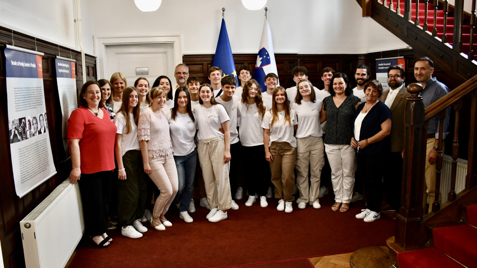 Državna sekretarka z maturanti in njihovimi spremljevalci. Stojijo, za njimi slovenska in evropska zastava.