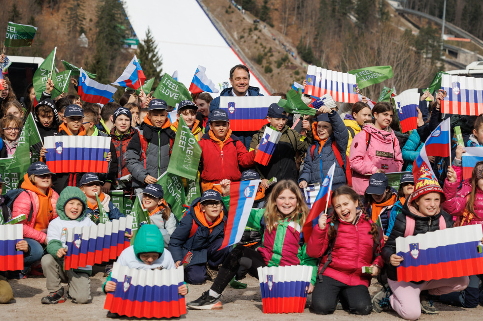 Minister in zamejski otroki držijo zastavice slovenske in i feel Slovenia. Otroci so nasmejani.