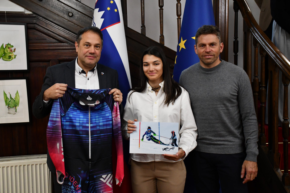 Matej Arčon v rokah drži dres, Caterina Sinigoi v rokah drži podpisano fotografijo in Aleš Sever. Za njimi slovenska in evropska zastava.